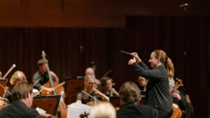 Olivia Clarke leads an orchestra during a performance. Musicians play various string instruments, and sheet music is visible on their stands. The setting is a concert hall with warm lighting.
