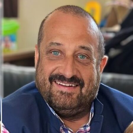 A smiling man with a beard and blue eyes, wearing a dark jacket over a checked shirt, sits indoors on a sofa. The blurred background hints at household items—perhaps reflecting his involvement in the Community Advisory Group.