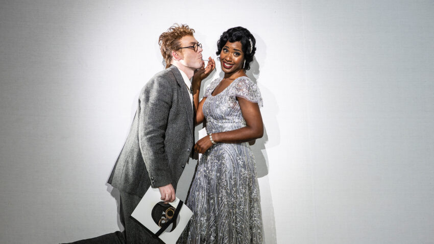 Two people pose playfully against a white background. One person in a grey suit leans in as if whispering, holding a mask and papers. The other, in a sparkly silver gown, smiles whilst looking at the camera.
