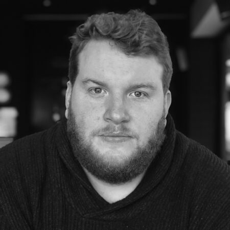 Samuel Downes, a man with curly hair and a beard, looks directly at the camera in a dark jumper. The softly blurred background and black-and-white tones add depth to the portrait.