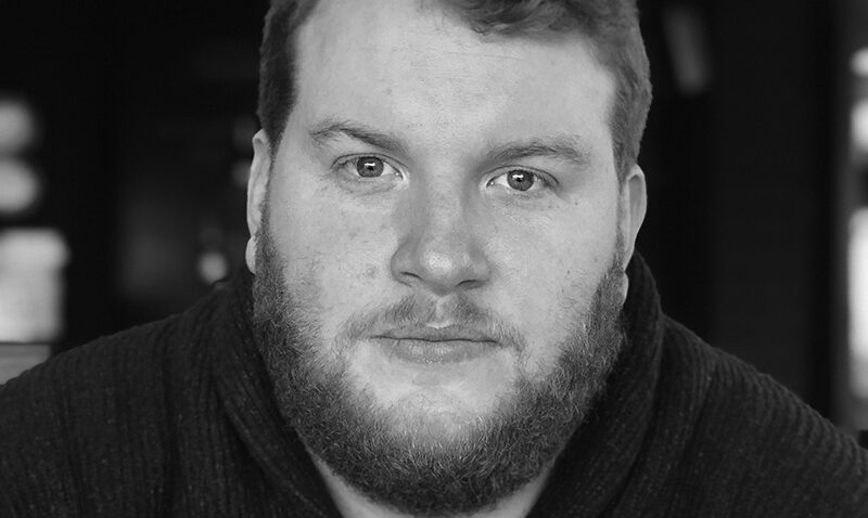 Samuel Downes, a man with curly hair and a beard, looks directly at the camera in a dark jumper. The softly blurred background and black-and-white tones add depth to the portrait.