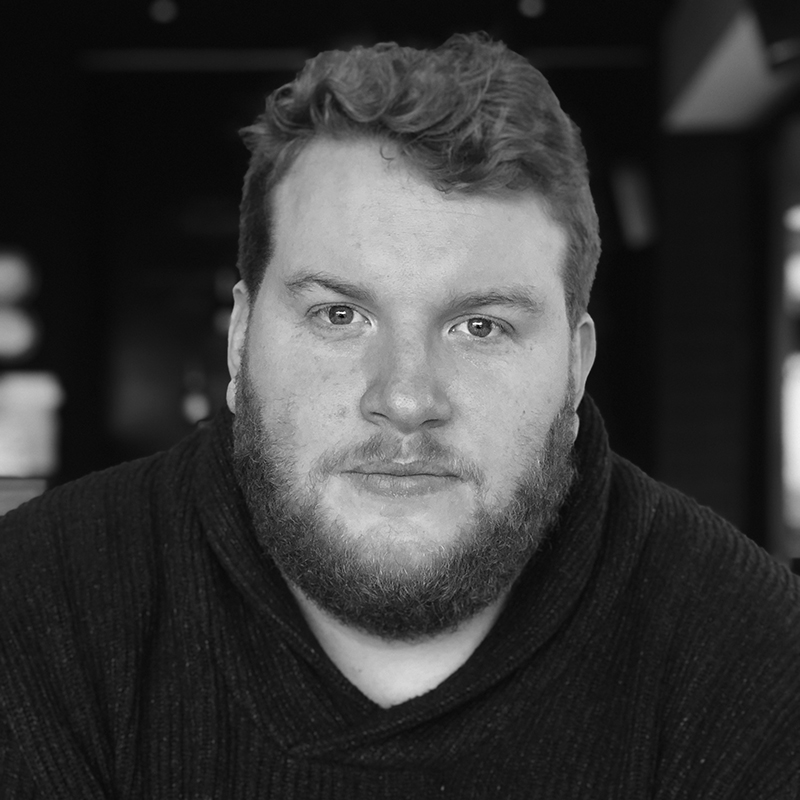 Samuel Downes, a man with curly hair and a beard, looks directly at the camera in a dark jumper. The softly blurred background and black-and-white tones add depth to the portrait.