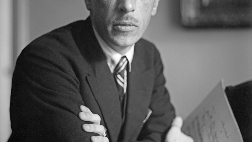A serious man in a suit sits with arms folded, holding sheet music. He has short dark hair, a thin moustache, and looks directly at the camera—resembling composer Igor Stravinsky. The blurred background shows part of a picture frame on the wall.