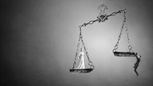 A black and white image of a balance scale, with Lady Justice on one side and a small person hanging from the other, evokes the weight of Trial By Jury against a plain gradient background.