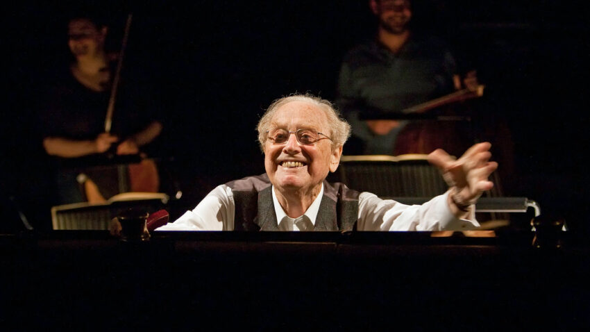 Sir Charles Mackerras with glasses smiles and gestures energetically whilst seated at a piano, as two musicians play classical music on string instruments in the dimly lit background.