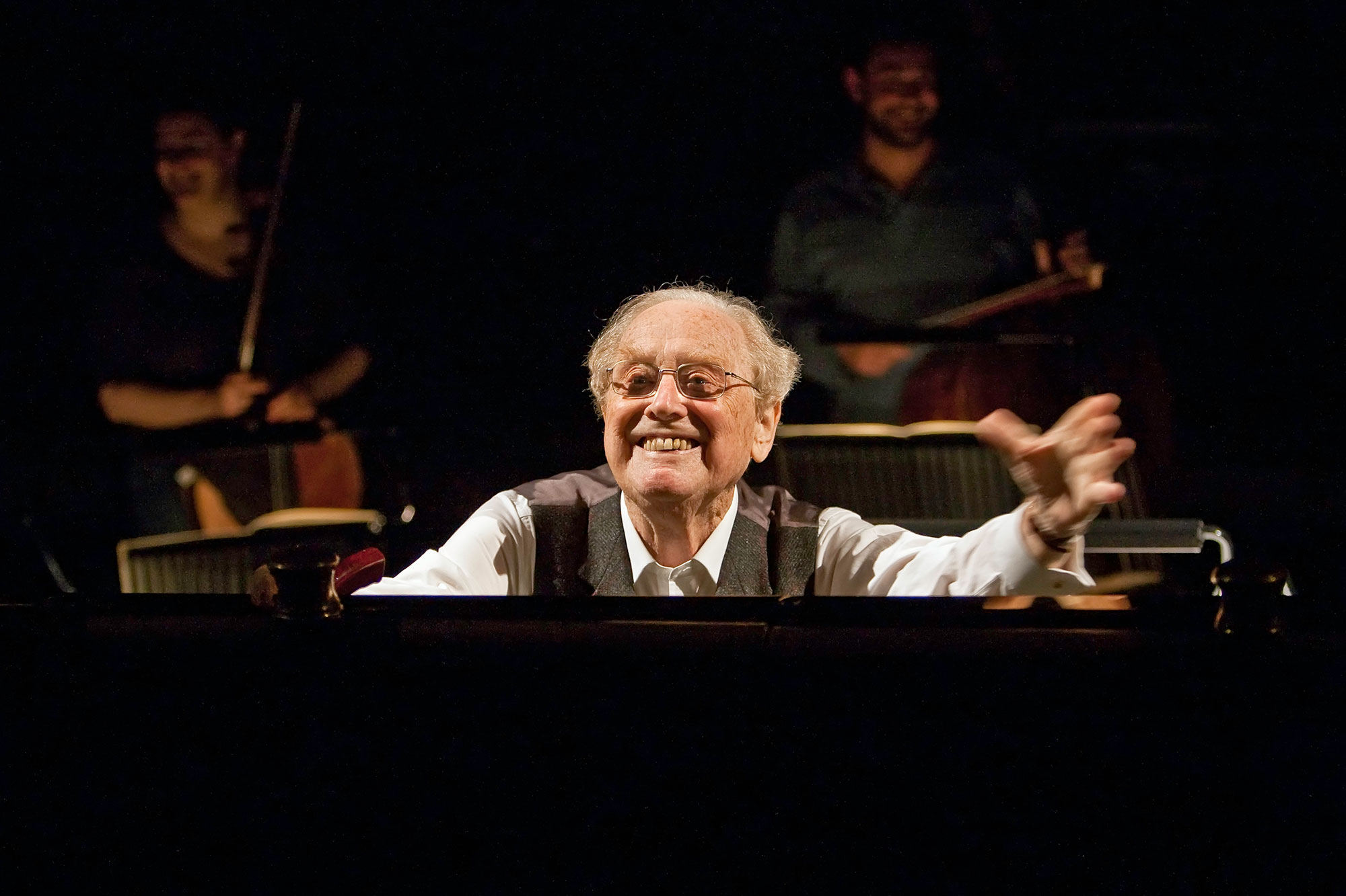 Sir Charles Mackerras with glasses smiles and gestures energetically whilst seated at a piano, as two musicians play classical music on string instruments in the dimly lit background.
