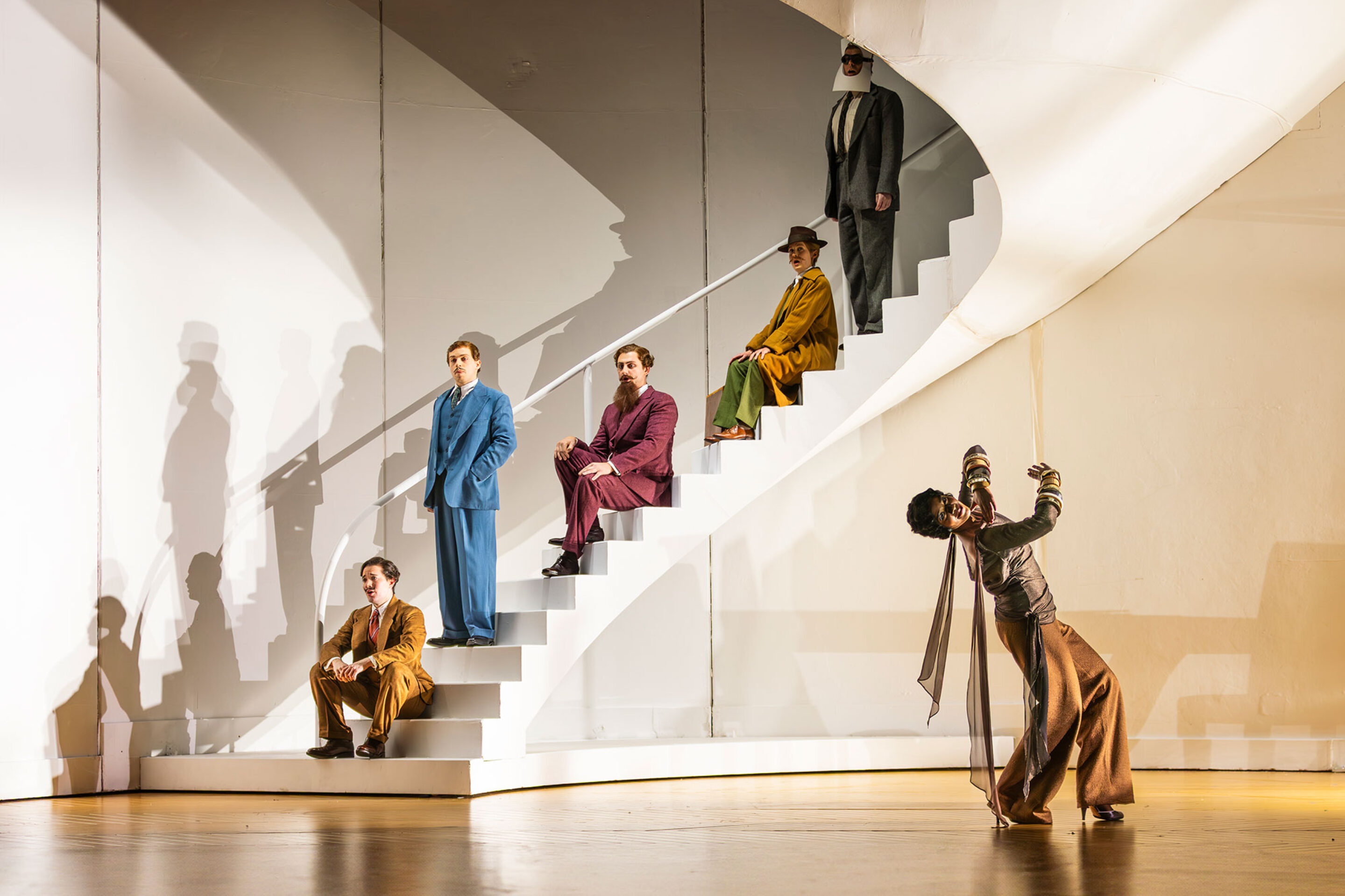 Five people in colourful, vintage-style outfits pose on a white, modern spiral staircase; one person stands dramatically at the base, while the others are positioned on different steps under dramatic lighting.