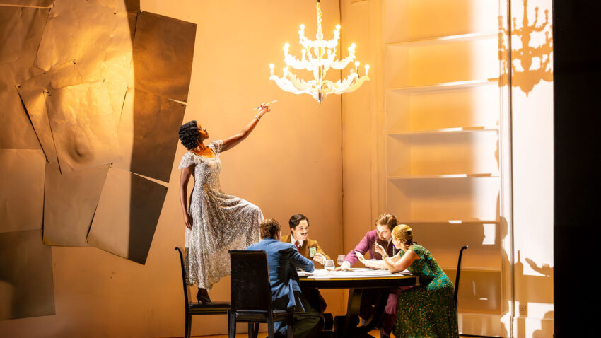 A woman in a floral dress stands on a dining table reaching towards a lit chandelier, while four people sit around the table watching her in a brightly lit, modern room with minimalist decor.