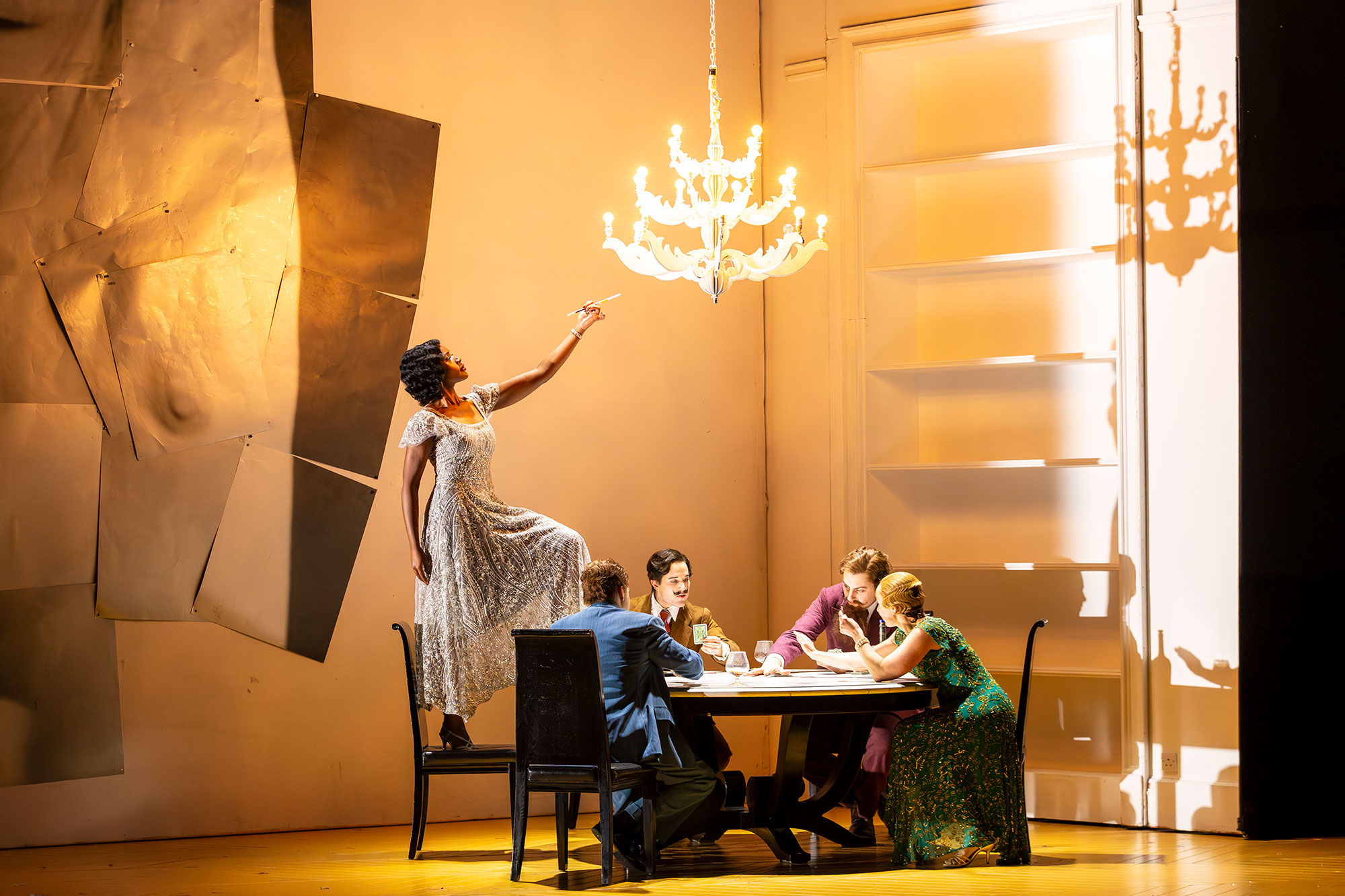 A woman in a floral dress stands on a dining table reaching towards a lit chandelier, while four people sit around the table watching her in a brightly lit, modern room with minimalist decor.