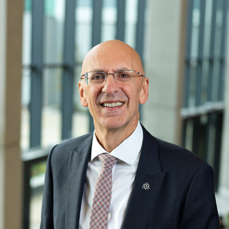 Professor Malcolm Press, a bald man wearing glasses, a dark suit, white shirt, and patterned tie, smiles while standing indoors in front of tall windows with vertical lines.