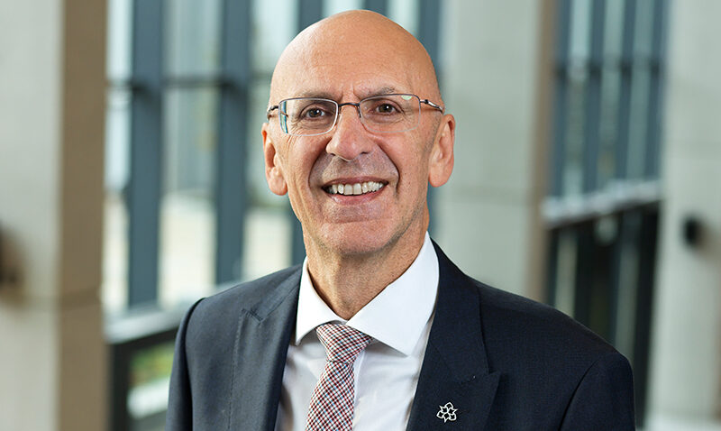 Professor Malcolm Press, a bald man wearing glasses, a dark suit, white shirt, and patterned tie, smiles while standing indoors in front of tall windows with vertical lines.