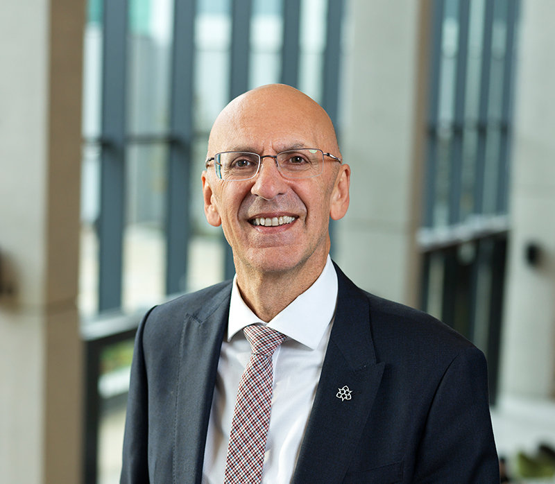 Professor Malcolm Press, a bald man wearing glasses, a dark suit, white shirt, and patterned tie, smiles while standing indoors in front of tall windows with vertical lines.
