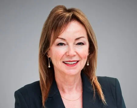 Vanda Murray, a woman with straight, light brown hair wearing a dark blazer and pearl drop earrings, smiles in front of a plain grey background.