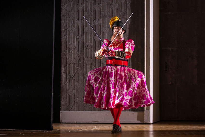 A performer in a bright pink floral dress, red tights, and black shoes stands on stage, wearing a helmet with a yellow plume and holding two crossed batons. The background is dark and textured.