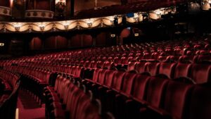 Rows of empty red velvet seats in a dimly lit, ornate theatre auditorium, facing a stage. The upper balcony with decorative railings and lights is visible in the background.