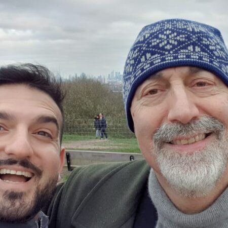 Two men from the Community Advisory Group smile and take a selfie outdoors on a cloudy day, with trees, grass, and a cityscape in the background. One wears a patterned blue winter hat, whilst the other has dark hair and a beard.