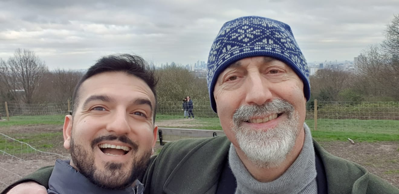 Two men from the Community Advisory Group smile and take a selfie outdoors on a cloudy day, with trees, grass, and a cityscape in the background. One wears a patterned blue winter hat, whilst the other has dark hair and a beard.