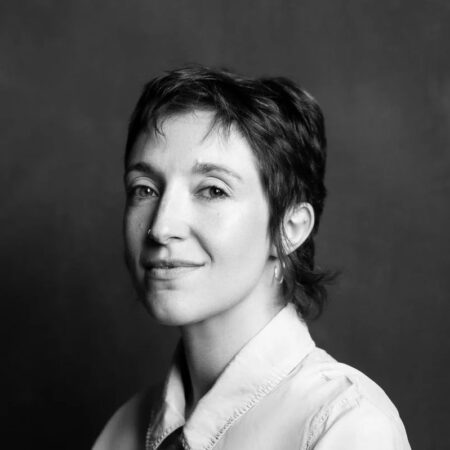 Black and white portrait of a Ruth Knight with short, dark hair, wearing a light-coloured collared shirt and hoop earrings, looking slightly to the side with a calm expression against a plain background.