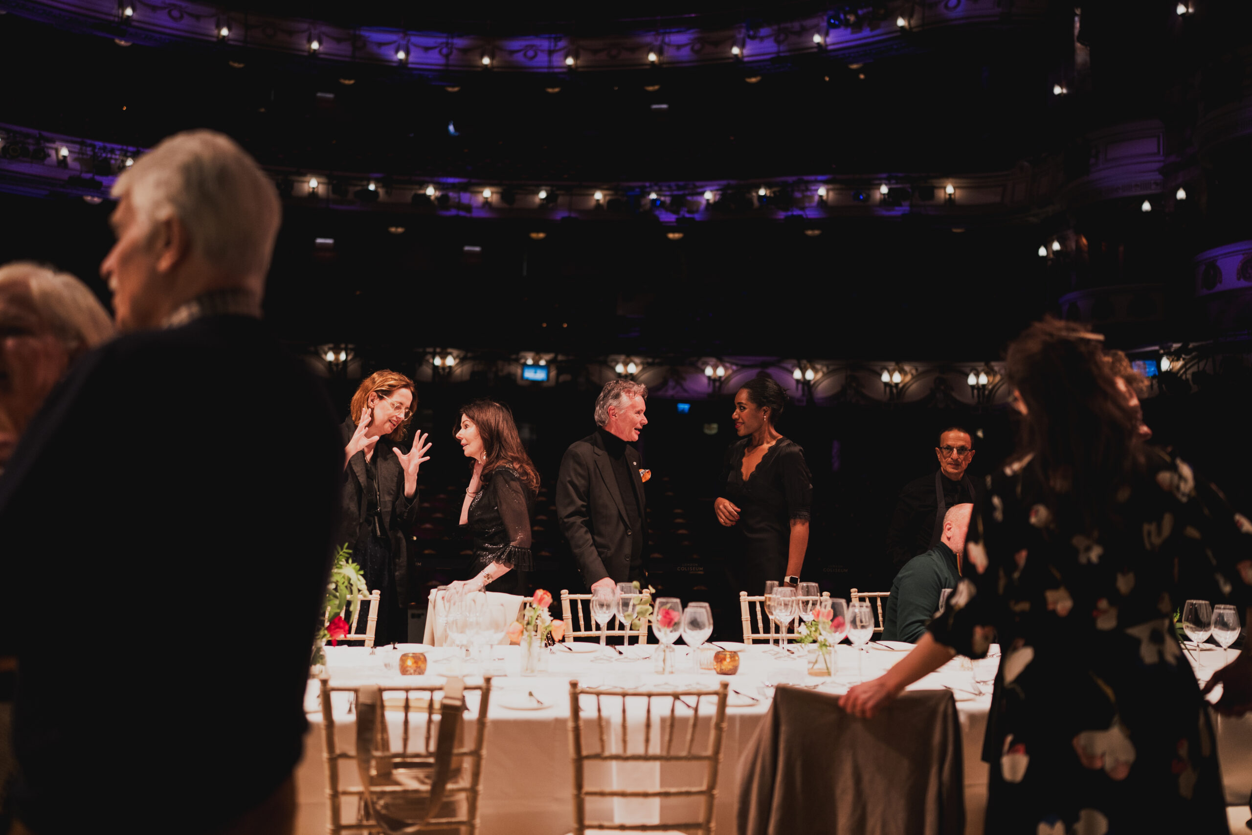 People dressed in semi-formal attire stand and converse around a long dining table set with glasses and plates, on a stage with empty theatre seats in the background and soft, purple lighting.