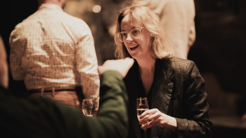 A woman with glasses and shoulder-length blonde hair smiles while holding a glass, engaging in conversation at a social gathering. Other people are visible in the softly lit, blurred background.