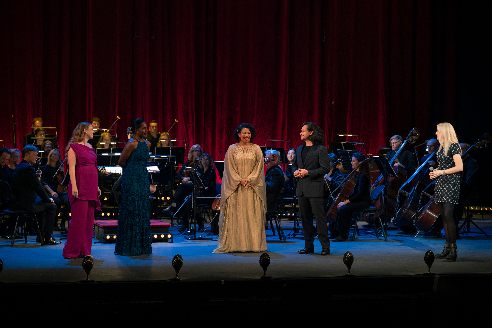 Four performers stand on stage in front of a seated orchestra, with red curtains behind them. Three women and one man appear to be singing or speaking, dressed in formal and elegant attire.