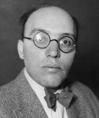 A black-and-white portrait of a man with round glasses, wearing a bow tie, a suit jacket, and a serious expression, against a plain dark background.