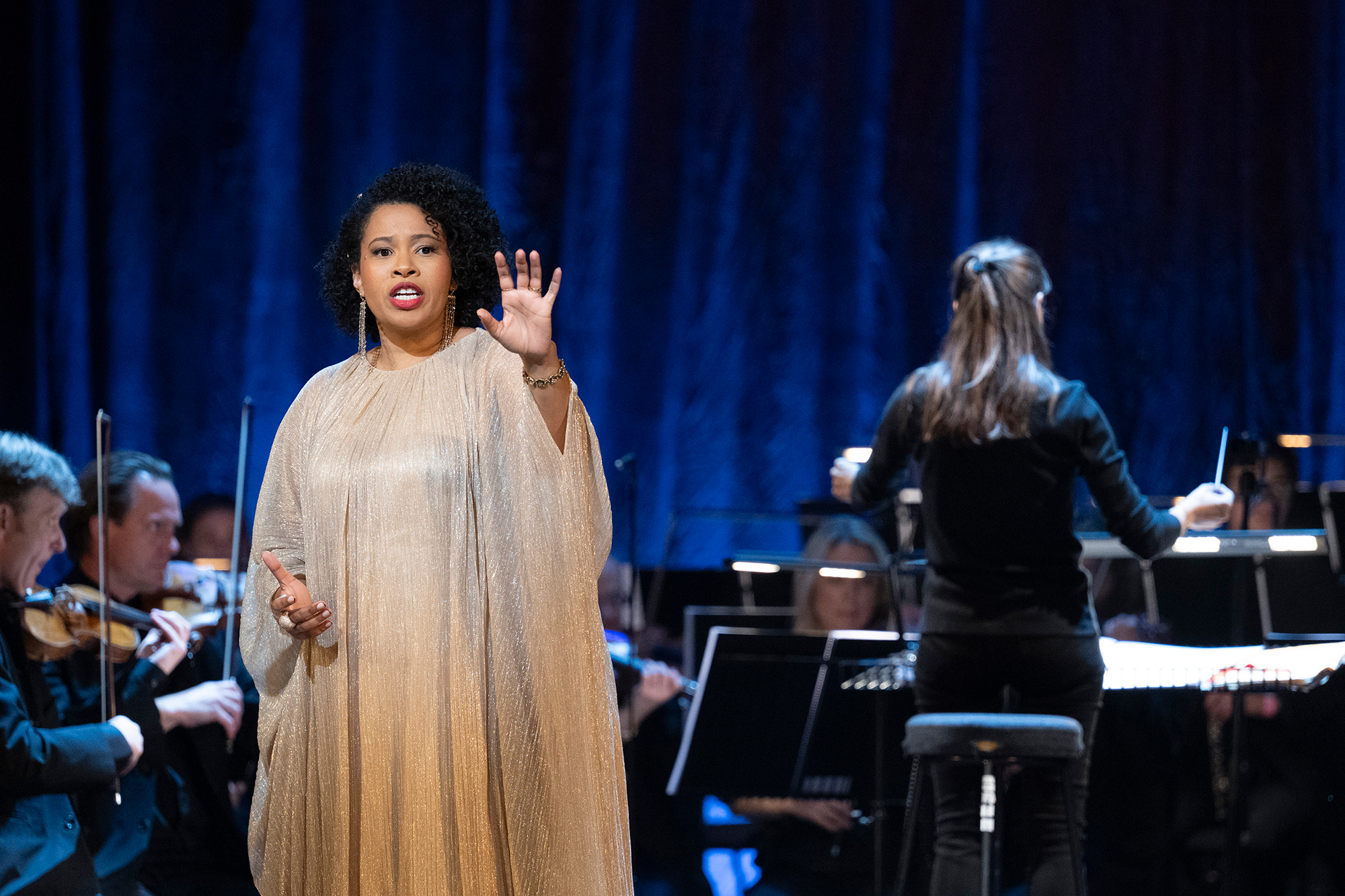 A female soloist in a flowing gold dress sings on stage with an orchestra behind her; a conductor with long hair stands facing the musicians, leading the performance against a dark blue backdrop.