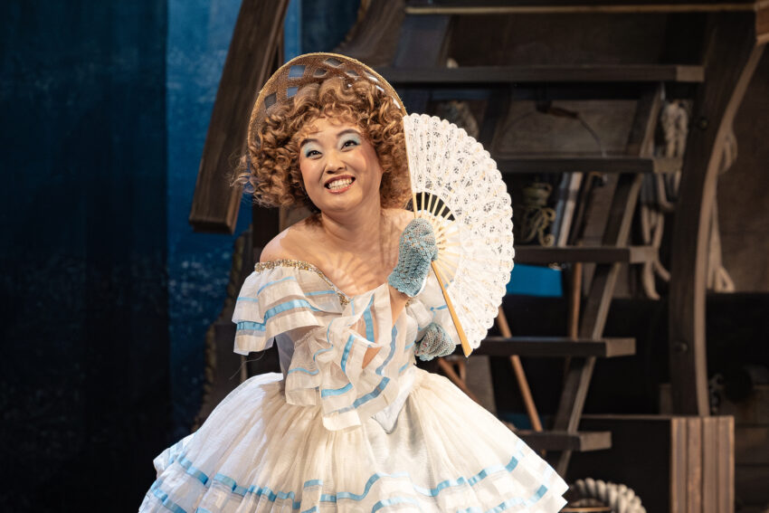 A smiling woman in a curly wig and frilly white dress with blue accents holds a lace fan and wears blue gloves, standing in front of a wooden staircase on a theatrical stage.