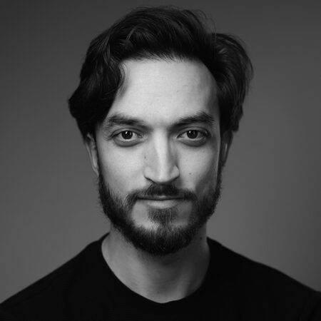 Black and white portrait of a young man with wavy hair, a beard, and moustache, wearing a dark shirt. He is looking directly at the camera with a slight smile against a plain background.