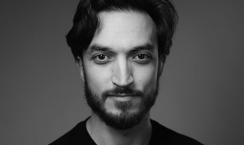 Black and white portrait of a young man with wavy hair, a beard, and moustache, wearing a dark shirt. He is looking directly at the camera with a slight smile against a plain background.