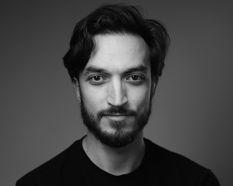 Black and white portrait of a young man with wavy hair, a beard, and moustache, wearing a dark shirt. He is looking directly at the camera with a slight smile against a plain background.