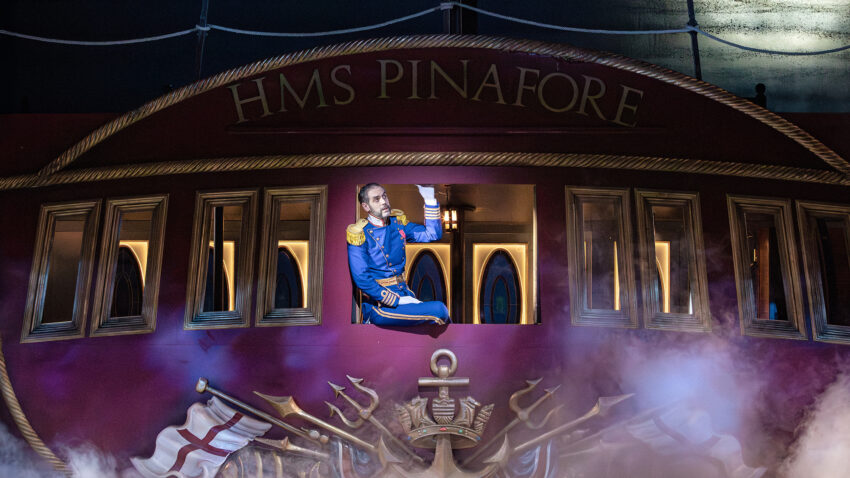 A performer in a blue naval uniform leans out of a window on a ship set labelled HMS Pinafore, surrounded by stage smoke and nautical decorations.