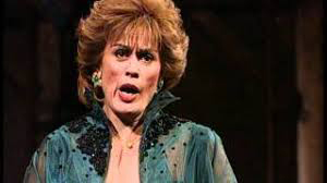 A woman with short, styled hair and bold make-up wears a teal, sheer, embellished outfit and large green earrings, looking ahead with an expressive facial expression—capturing the drama of a musical journey worthy of Mozart.