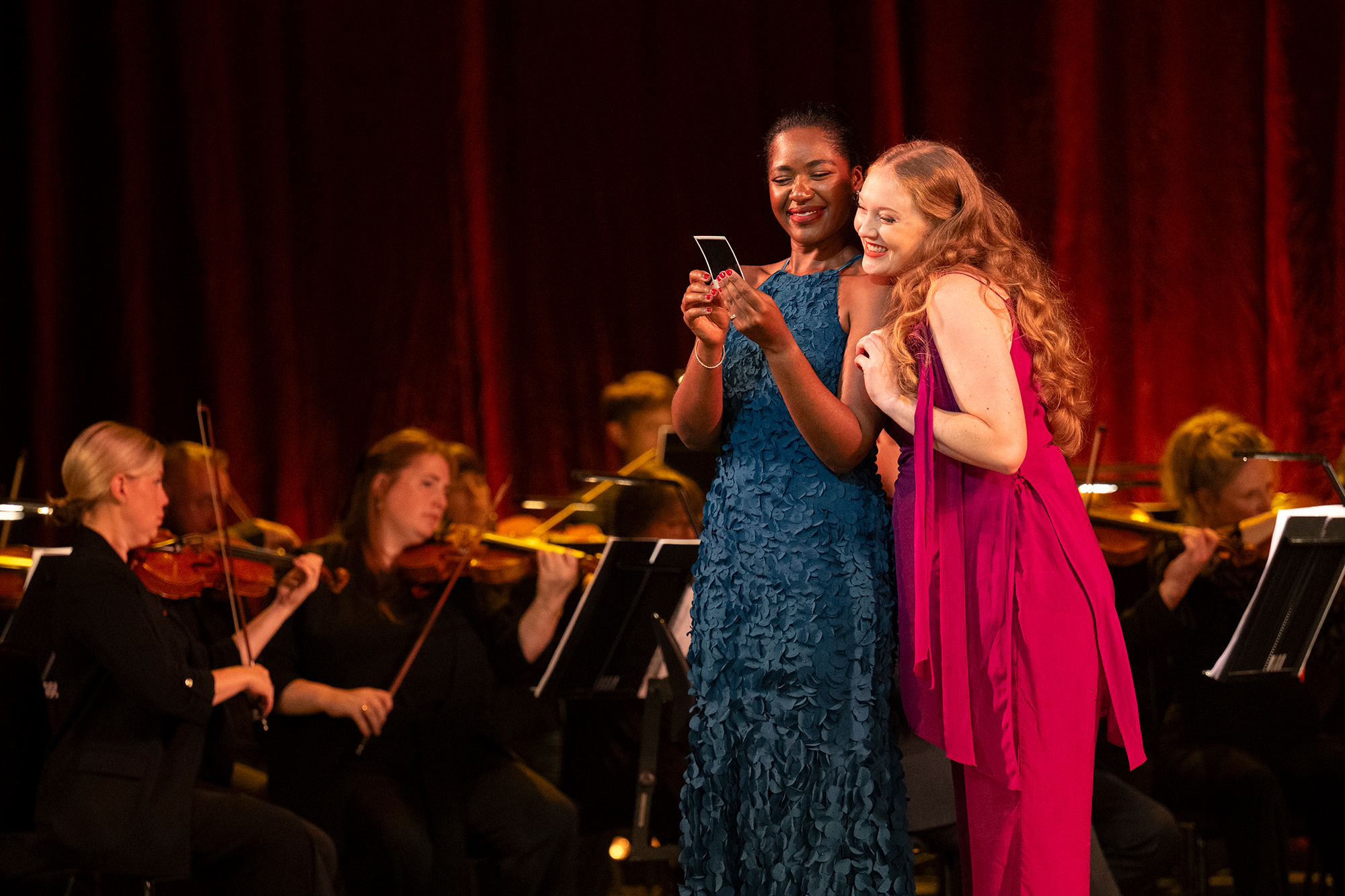 Two women in elegant dresses stand on stage, smiling and looking at a mobile, with an orchestra of violinists playing behind them against a red curtain backdrop.