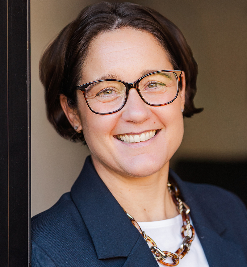 Nicola Cardillo-Zallo, a person with short brown hair and glasses smiles at the camera, wearing a dark blazer, white top, and a chunky chain necklace. The background is softly blurred.