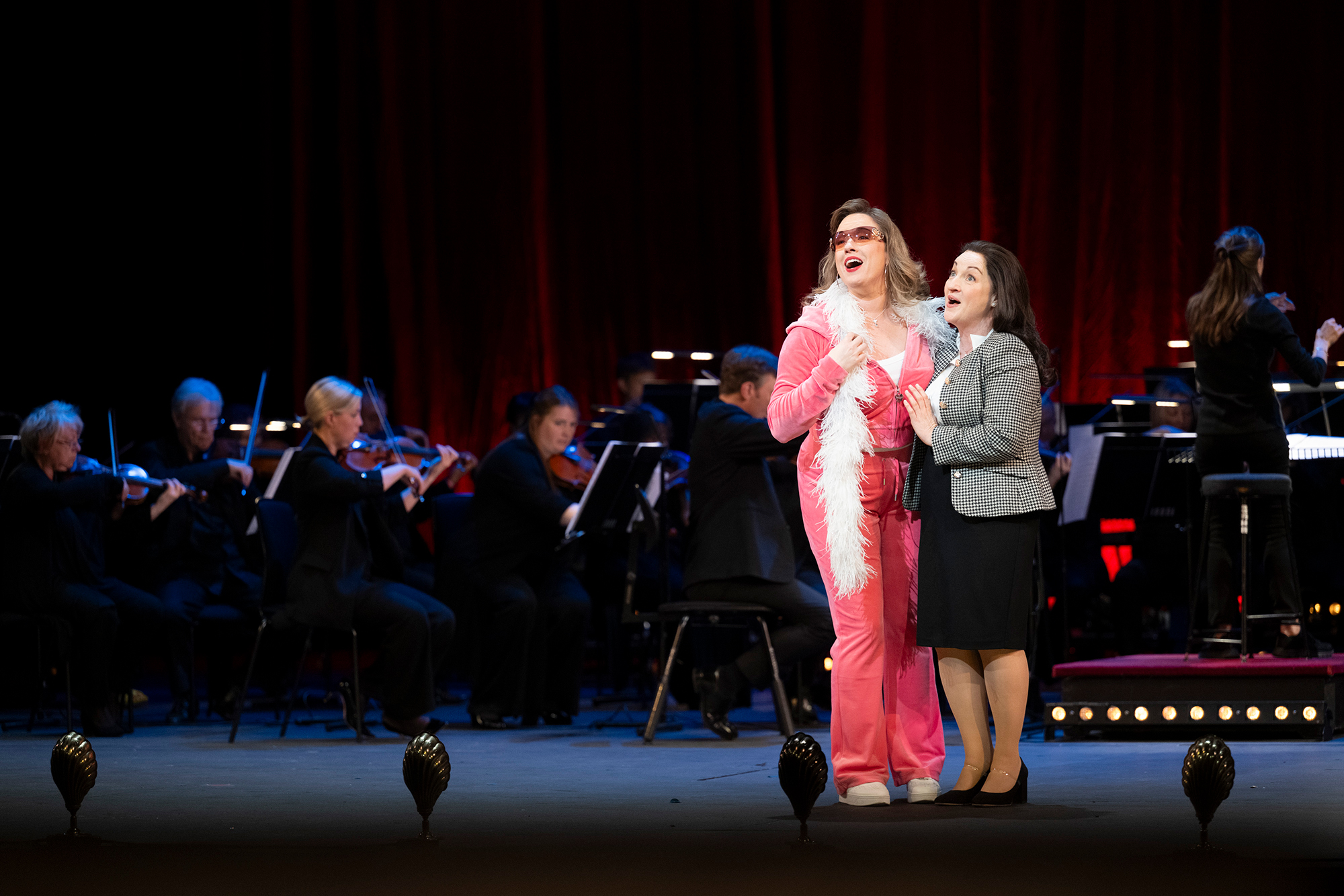 Two women sing passionately on stage in front of an orchestra; one wears a pink outfit with a feather boa, and the other wears a chequered blazer and skirt. Musicians play string instruments under soft stage lighting.