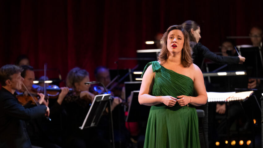 A woman in a green dress sings on stage in front of an orchestra, with musicians playing string instruments and a conductor directing them, all set against a dark red curtain backdrop.