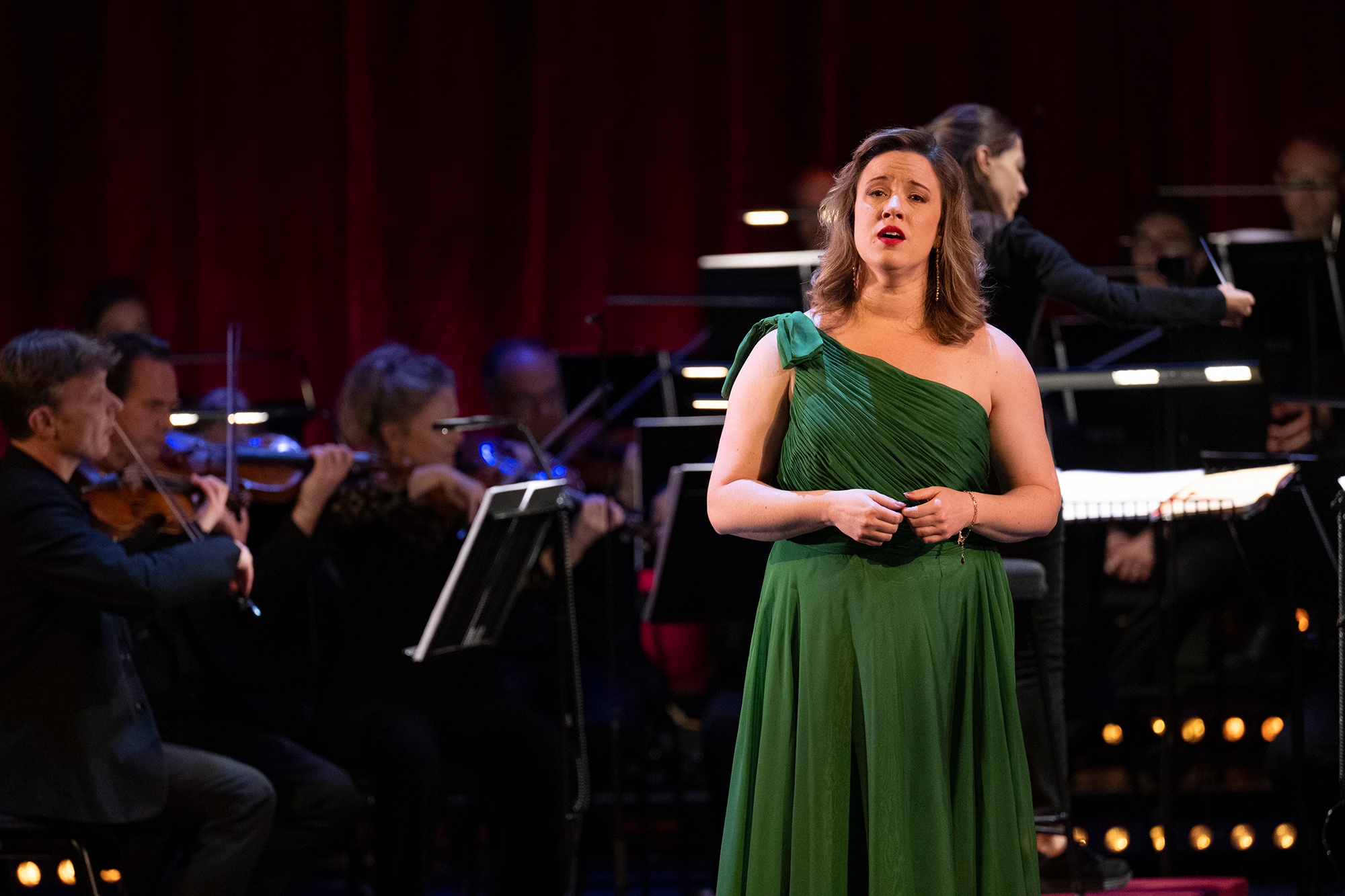 A woman in a green dress sings on stage in front of an orchestra, with musicians playing string instruments and a conductor directing them, all set against a dark red curtain backdrop.
