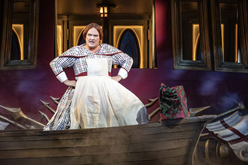 A person in a checked cape and white apron stands assertively on a wooden boat on stage, with dramatic lighting and an ornate red and gold background, evoking a theatrical performance.