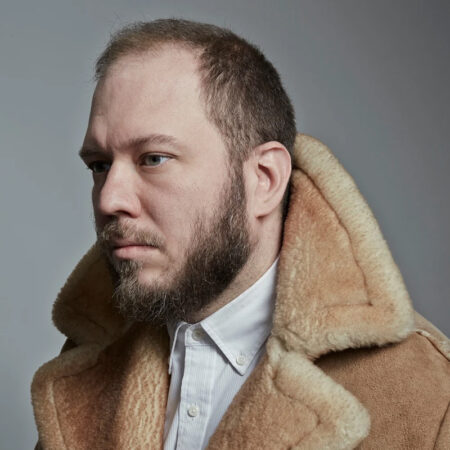 A bearded man with short hair, resembling Royce Vavrek, wears a tan shearling coat and a white collared shirt as he looks to the left against a plain grey background.