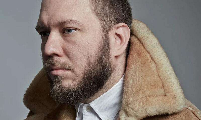 A bearded man with short hair, resembling Royce Vavrek, wears a tan shearling coat and a white collared shirt as he looks to the left against a plain grey background.