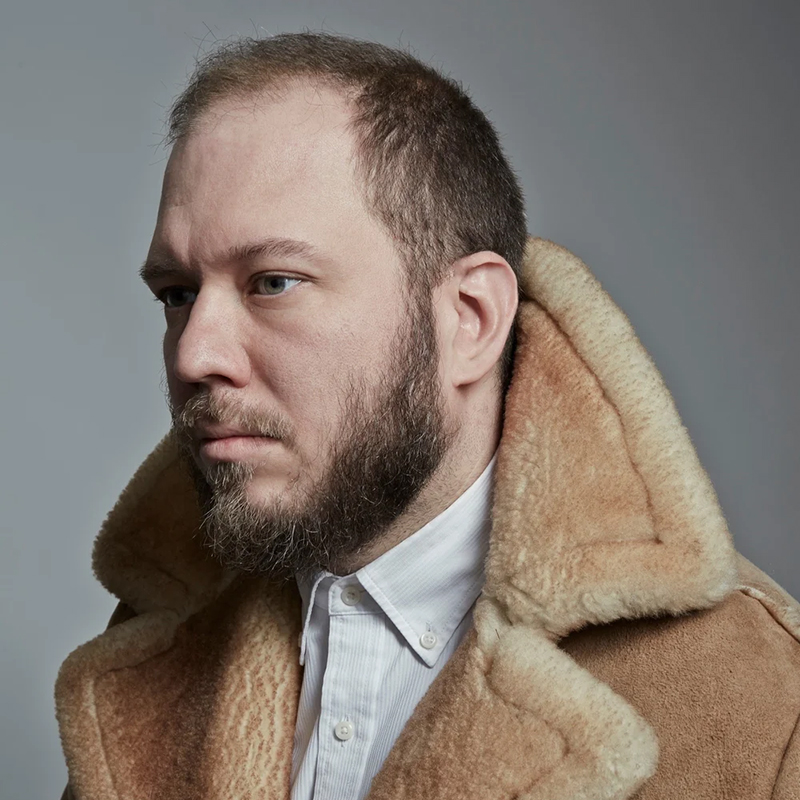 A bearded man with short hair, resembling Royce Vavrek, wears a tan shearling coat and a white collared shirt as he looks to the left against a plain grey background.