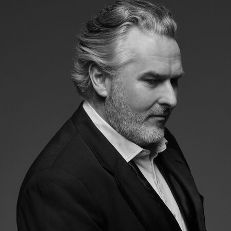A greyscale portrait of Simon O'Neill, a middle-aged man with grey hair and a beard, wearing a suit and dress shirt, looking down and to the right against a plain background.