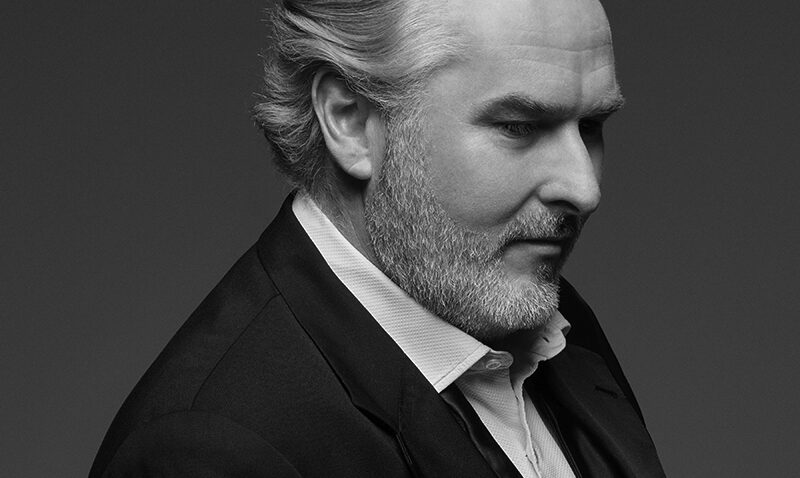 A greyscale portrait of Simon O'Neill, a middle-aged man with grey hair and a beard, wearing a suit and dress shirt, looking down and to the right against a plain background.