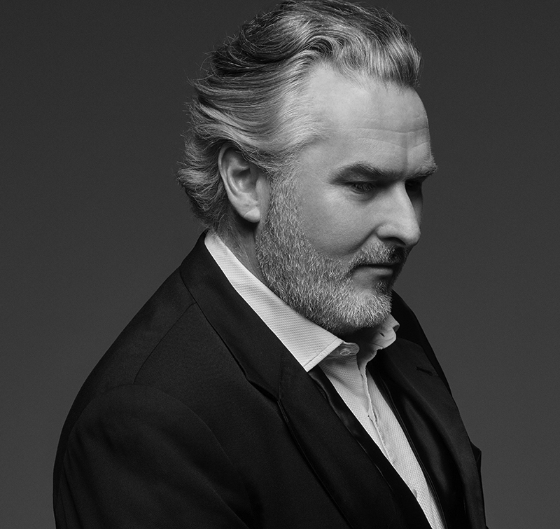 A greyscale portrait of Simon O'Neill, a middle-aged man with grey hair and a beard, wearing a suit and dress shirt, looking down and to the right against a plain background.