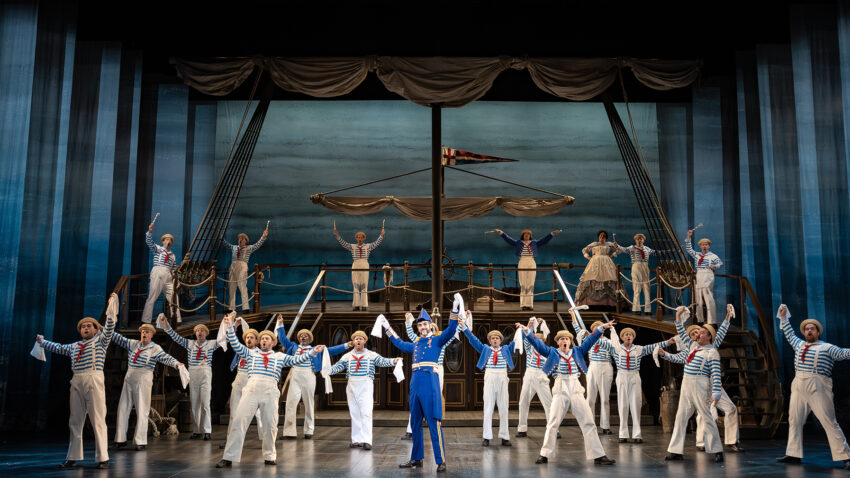 A large group of performers dressed as sailors in white trousers, striped shirts, and sailor hats sing and dance on a stage set resembling a ship, with one person in a blue uniform standing at the centre.