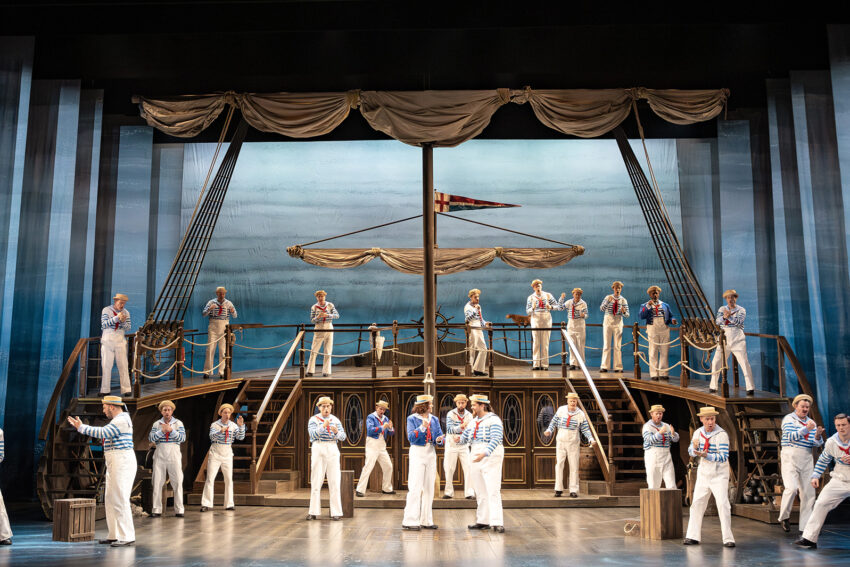 A large group of performers dressed as sailors stand and pose on a stylised ship set on stage, with blue striped shirts, white trousers, and nautical props, under dramatic lighting in a theatre production.