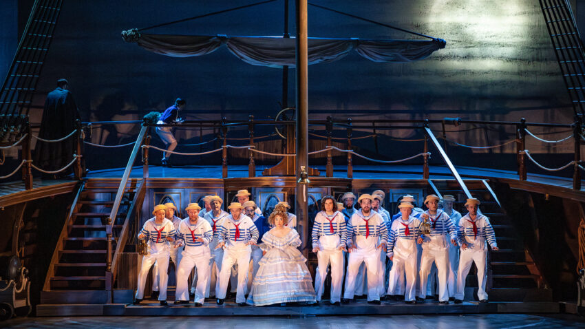 A group of performers in white sailor costumes and hats stand singing together on a ship-themed stage, with a woman in a white dress at the centre and dramatic lighting creating a maritime atmosphere.