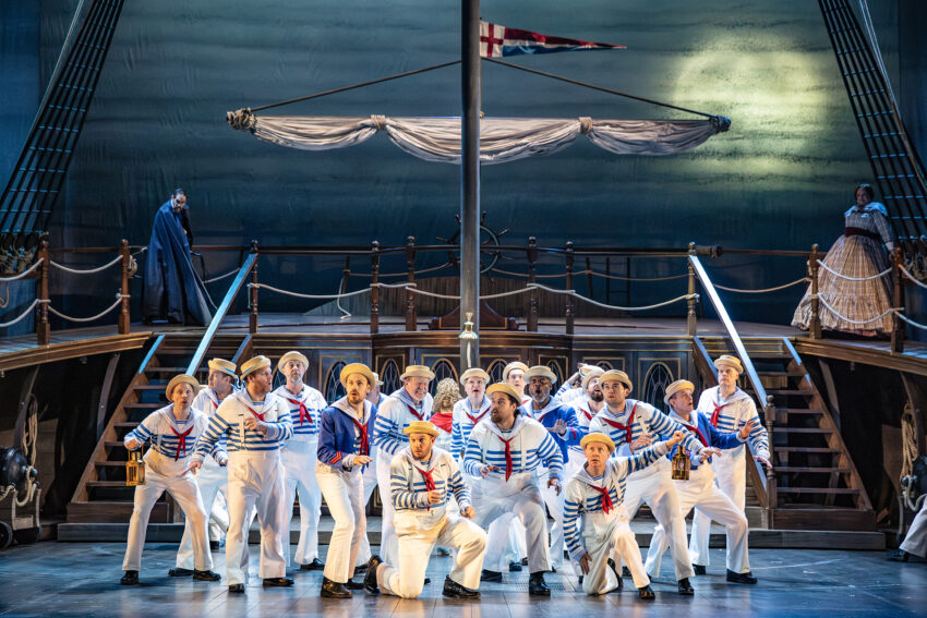 A group of performers dressed as sailors in white uniforms and hats pose together on a stage designed to look like a ship, with a large mast and rigging, and a dramatic nautical backdrop.