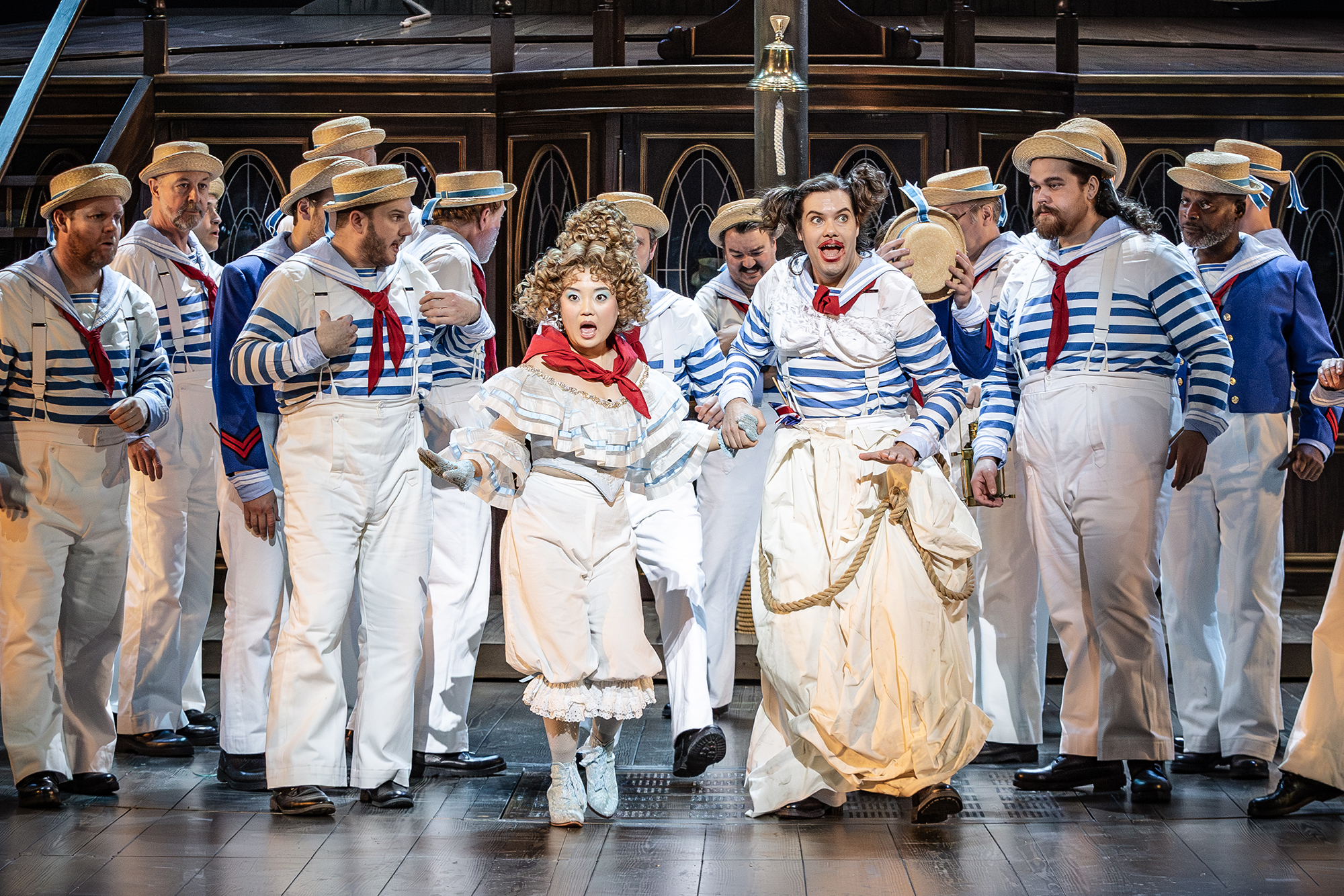 A theatrical scene with actors in sailor costumes and two performers in white dresses, one with curly blonde hair, centre stage. The background features wooden railings and decorative elements, evoking a nautical setting.